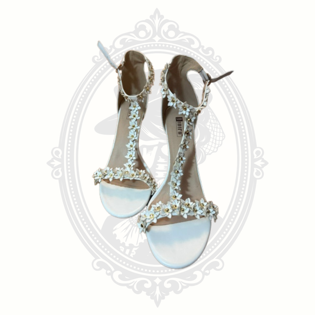 White floral appliqué high heel sandals with dimensional flower detailing and ankle straps displayed against a gothic-inspired background.