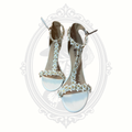 White floral appliqué high heel sandals with dimensional flower detailing and ankle straps displayed against a gothic-inspired background.