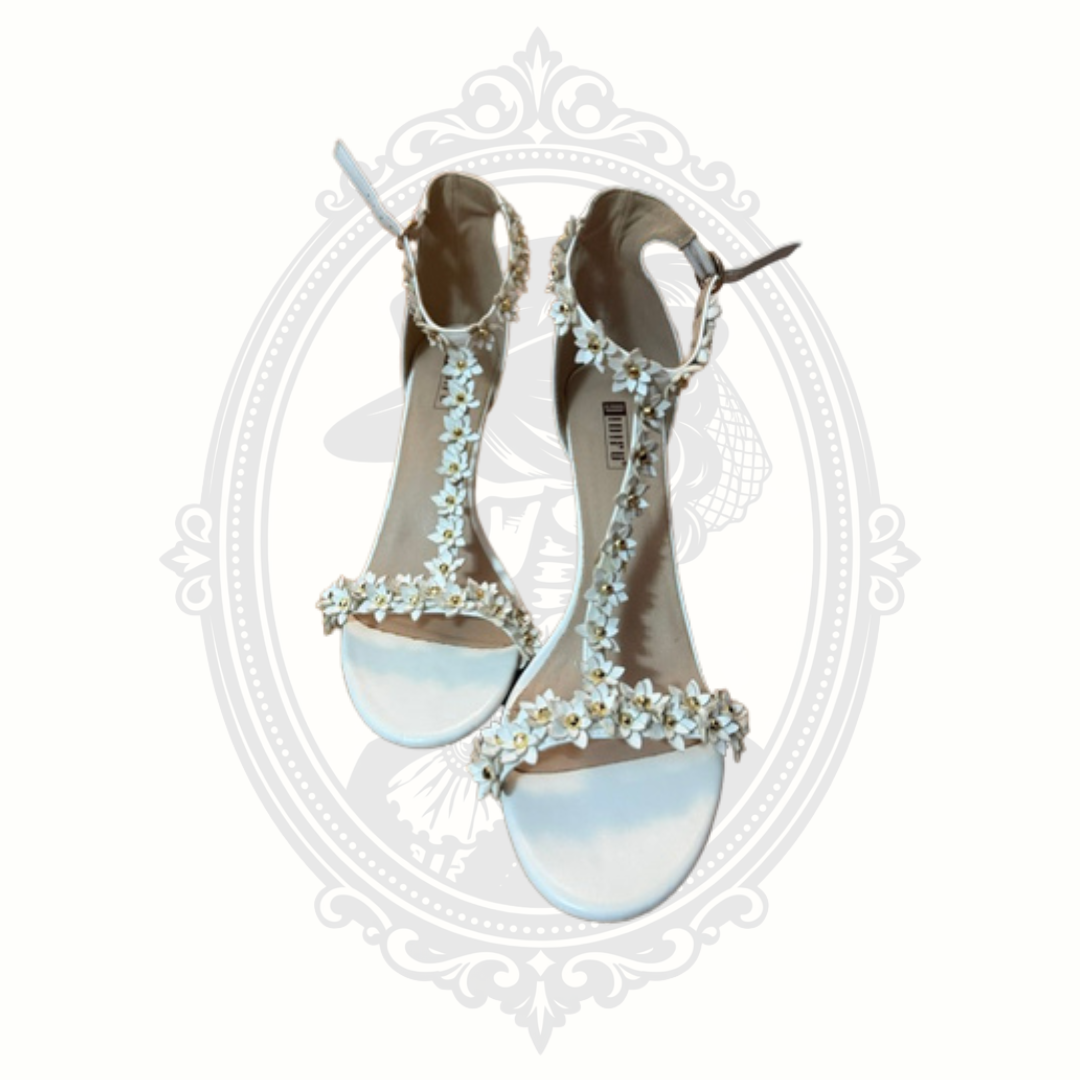 White floral appliqué high heel sandals with dimensional flower detailing and ankle straps displayed against a gothic-inspired background.