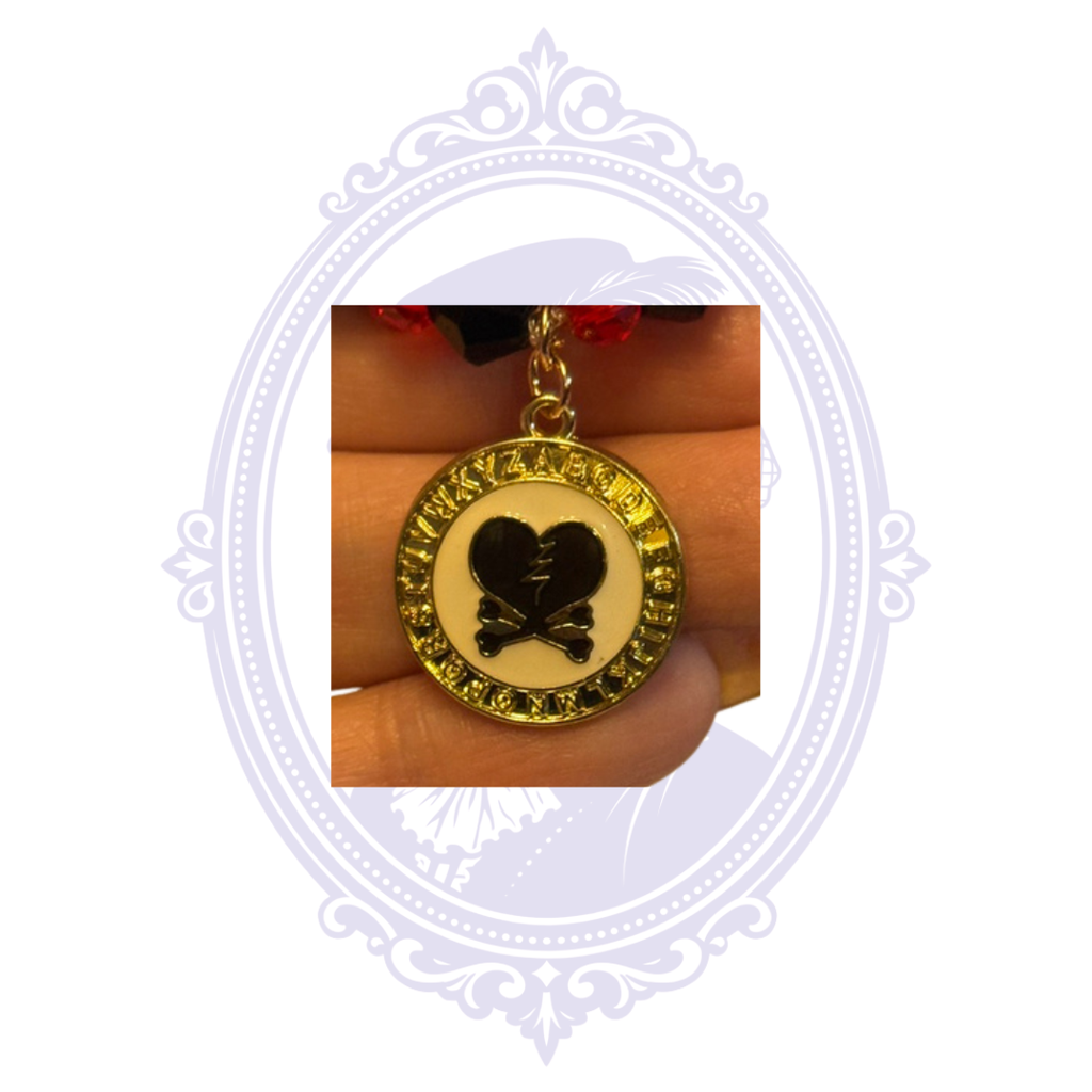 Detailed view of round gold-tone alphabet charm with black broken heart and crossbones design.