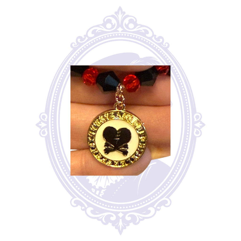 Close-up of red and black glass bead bracelet featuring a round gold-tone charm with black broken heart and crossbones design.