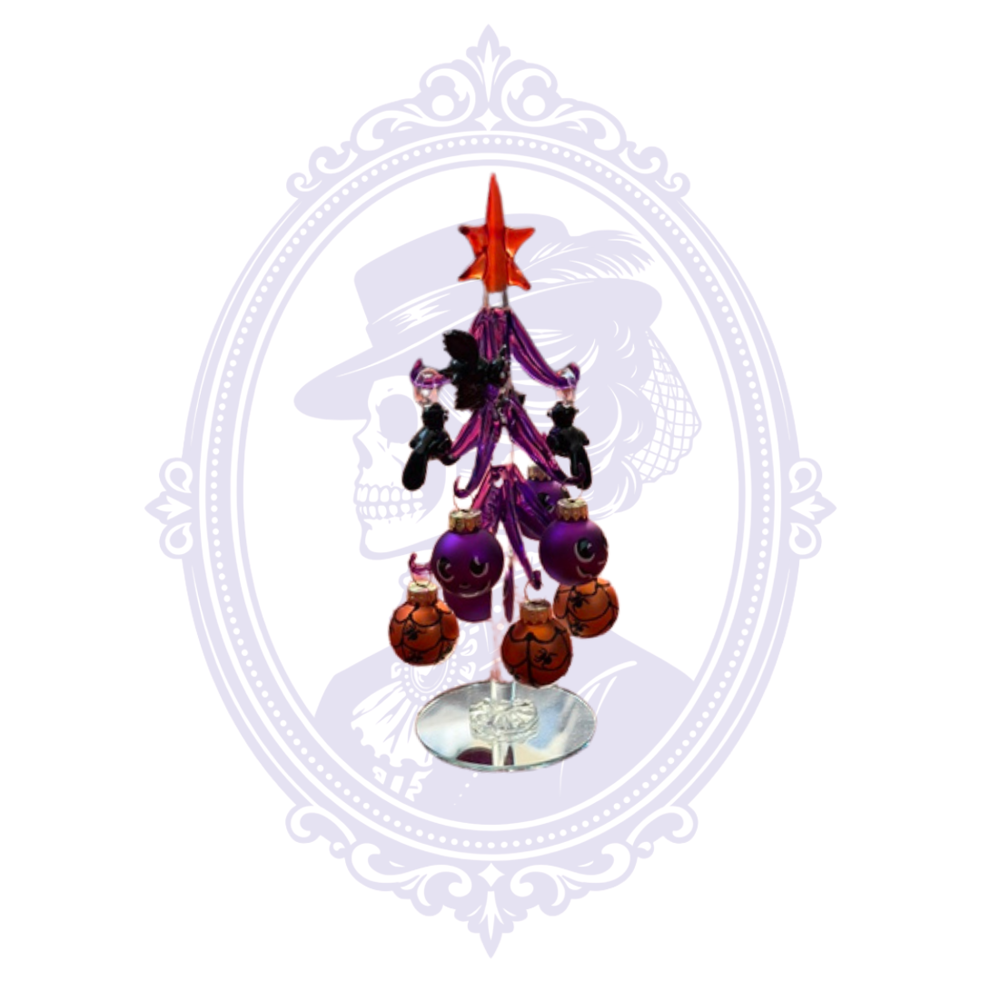 A small decorative Halloween tree with an orange star topper, purple branches, black ornaments, and orange baubles, displayed on a mirror base against a white background with a faint ornate frame.