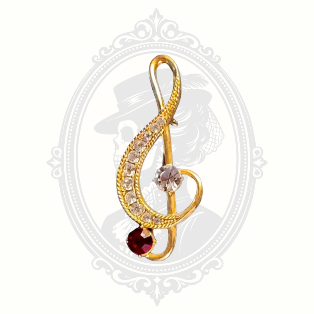 Gold treble clef brooch with rhinestones and a red gemstone accent, vintage style, displayed on a neutral background with a subtle Victorian cameo design.