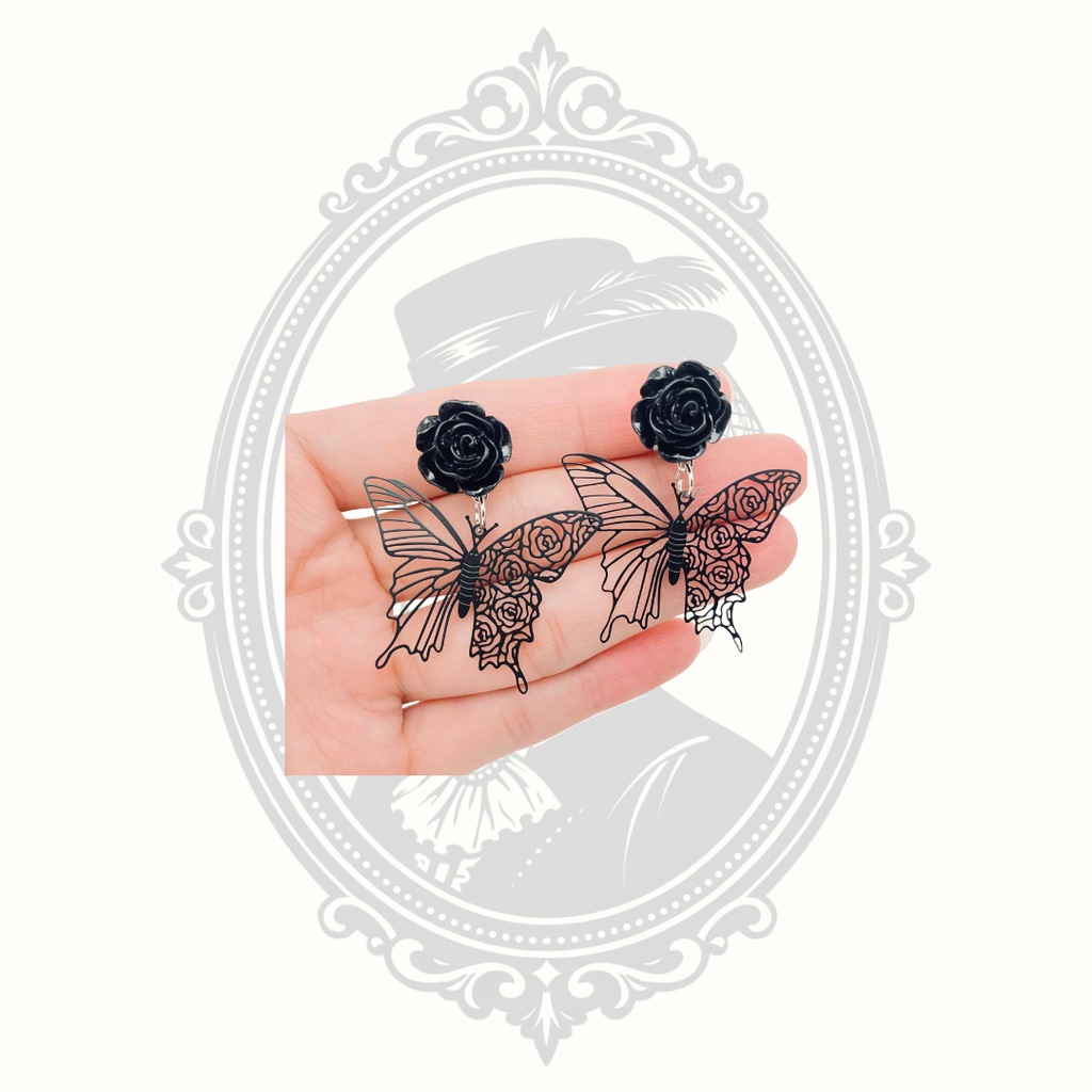 Black rose and butterfly stud earrings held in a hand, with a decorative gray vintage-style frame behind them.