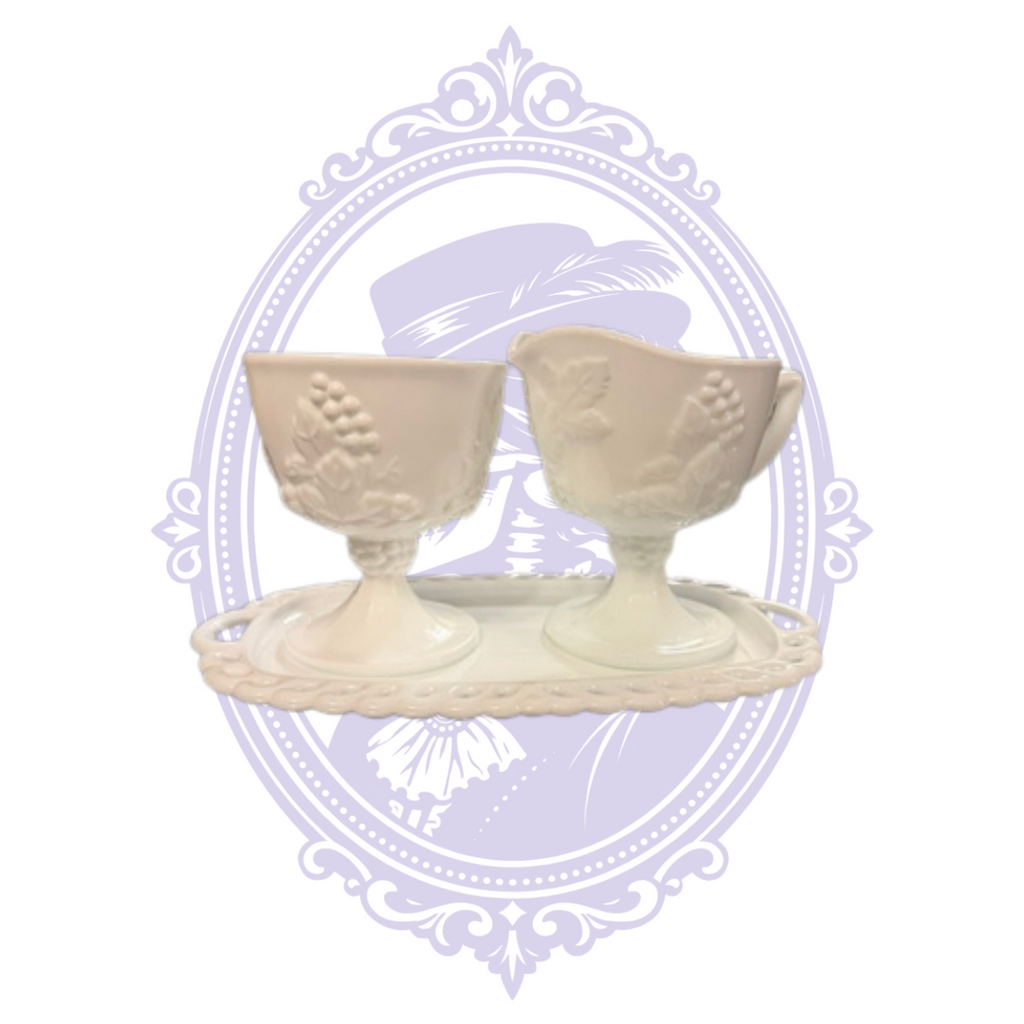 Vintage Colony Harvest white milk-glass three-piece serving set with a footed open sugar bowl and handled creamer, both decorated with embossed grape clusters and leaves. The pieces rest on their coordinating rectangular openwork undertray with lace-like edging, displayed against a pale lavender Mrs. Bones Curio Cupboard decorative background.