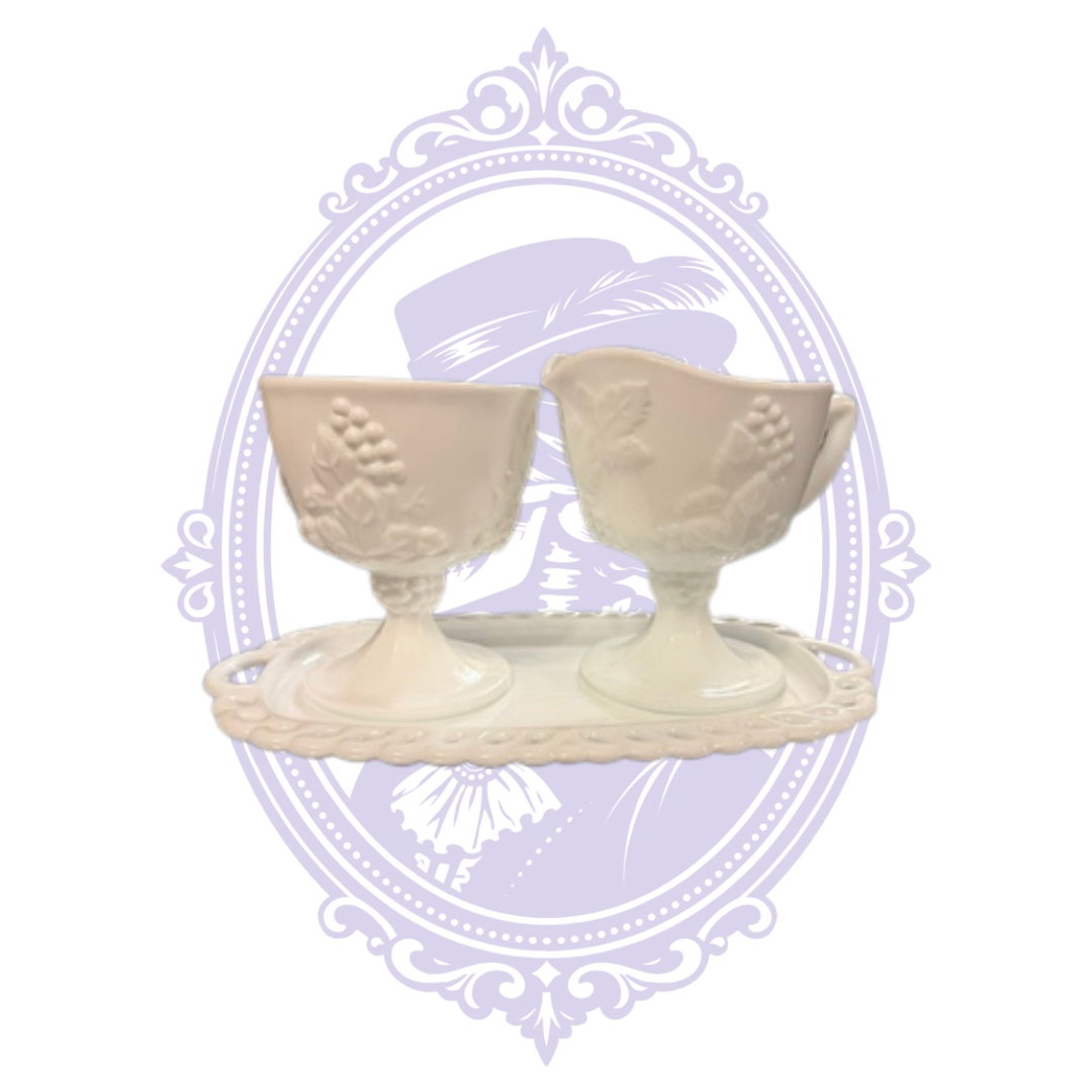 Vintage Colony Harvest white milk-glass three-piece serving set with a footed open sugar bowl and handled creamer, both decorated with embossed grape clusters and leaves. The pieces rest on their coordinating rectangular openwork undertray with lace-like edging, displayed against a pale lavender Mrs. Bones Curio Cupboard decorative background.