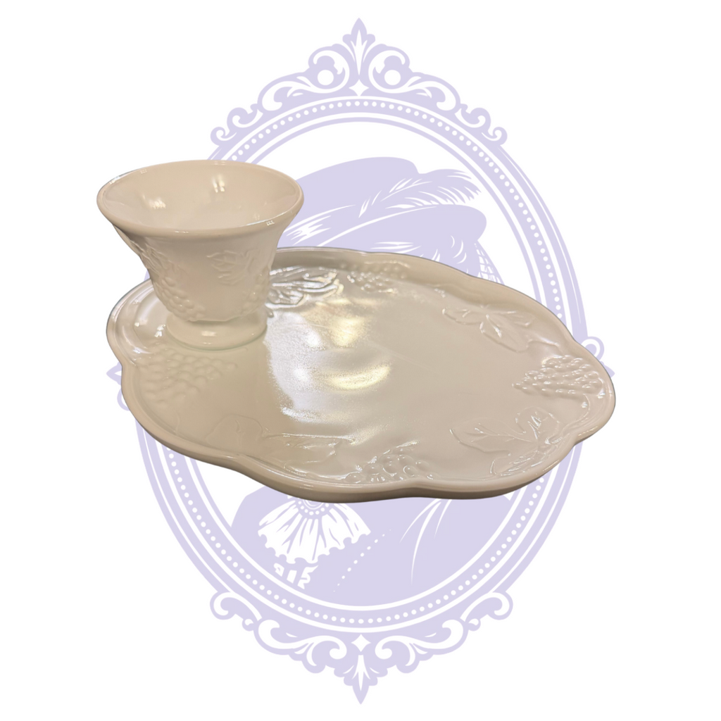 Vintage Indiana Glass Colony Harvest white milk-glass luncheon set featuring a pedestal-footed cup resting in the molded recess of a scalloped snack plate. Both pieces feature raised grape clusters and leaf designs, photographed against a pale lavender Mrs. Bones Curio Cupboard decorative background.