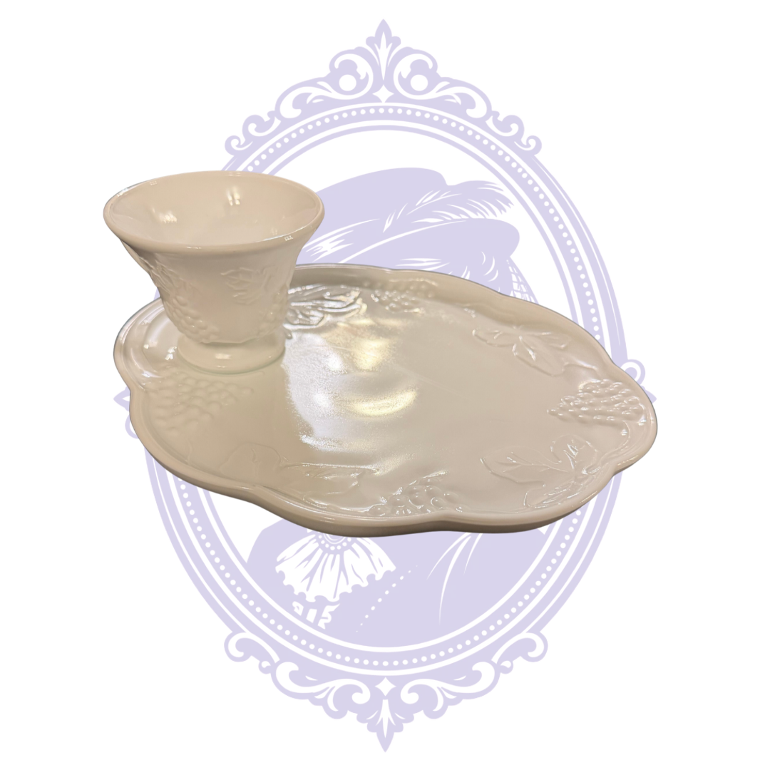 Vintage Indiana Glass Colony Harvest white milk-glass luncheon set featuring a pedestal-footed cup resting in the molded recess of a scalloped snack plate. Both pieces feature raised grape clusters and leaf designs, photographed against a pale lavender Mrs. Bones Curio Cupboard decorative background.
