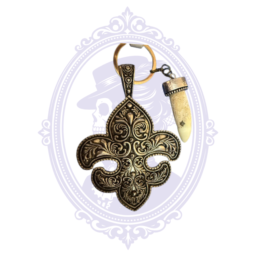 Close-up of a Louisiana-inspired keychain featuring an ornate antique-bronze-tone fleur-de-lis charm with intricate scrollwork and a genuine alligator tooth pendant in a decorative metal cap. Both hang from a gold-tone key ring. The keychain is displayed against a white background with a pale lavender Mrs. Bones Curio Cupboard skull logo and decorative oval frame.