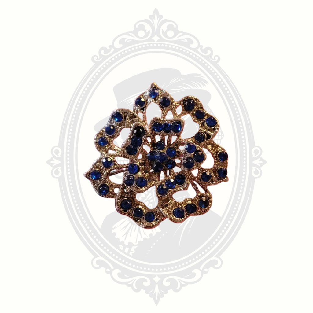 Vintage floral brooch with deep blue rhinestones set in gold-tone metal, featuring intricate openwork detailing and displayed on a soft neutral background with a faint Victorian cameo design.