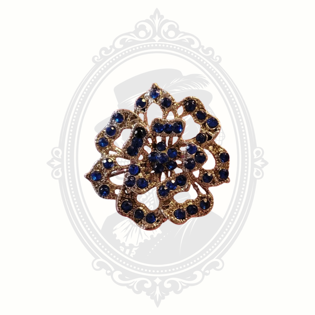 Vintage floral brooch with deep blue rhinestones set in gold-tone metal, featuring intricate openwork detailing and displayed on a soft neutral background with a faint Victorian cameo design.